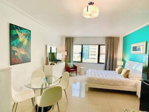 A hotel room with a bed, a desk with a chair, a TV, a painting, and a window with curtains.