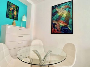 A dining area with a glass table and white chairs, a white cabinet with drawers, and two paintings on the wall.