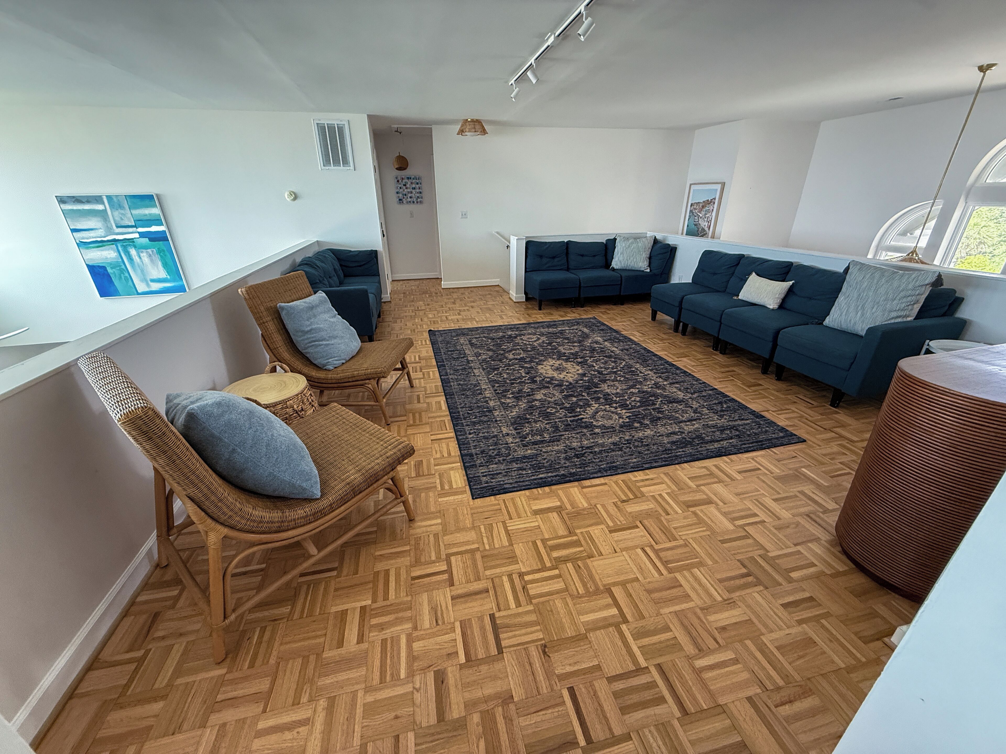 Een ruime woonkamer met een visgraatparketvloer, twee donkerblauwe banken, een rieten fauteuil en een groot donker vloerkleed.