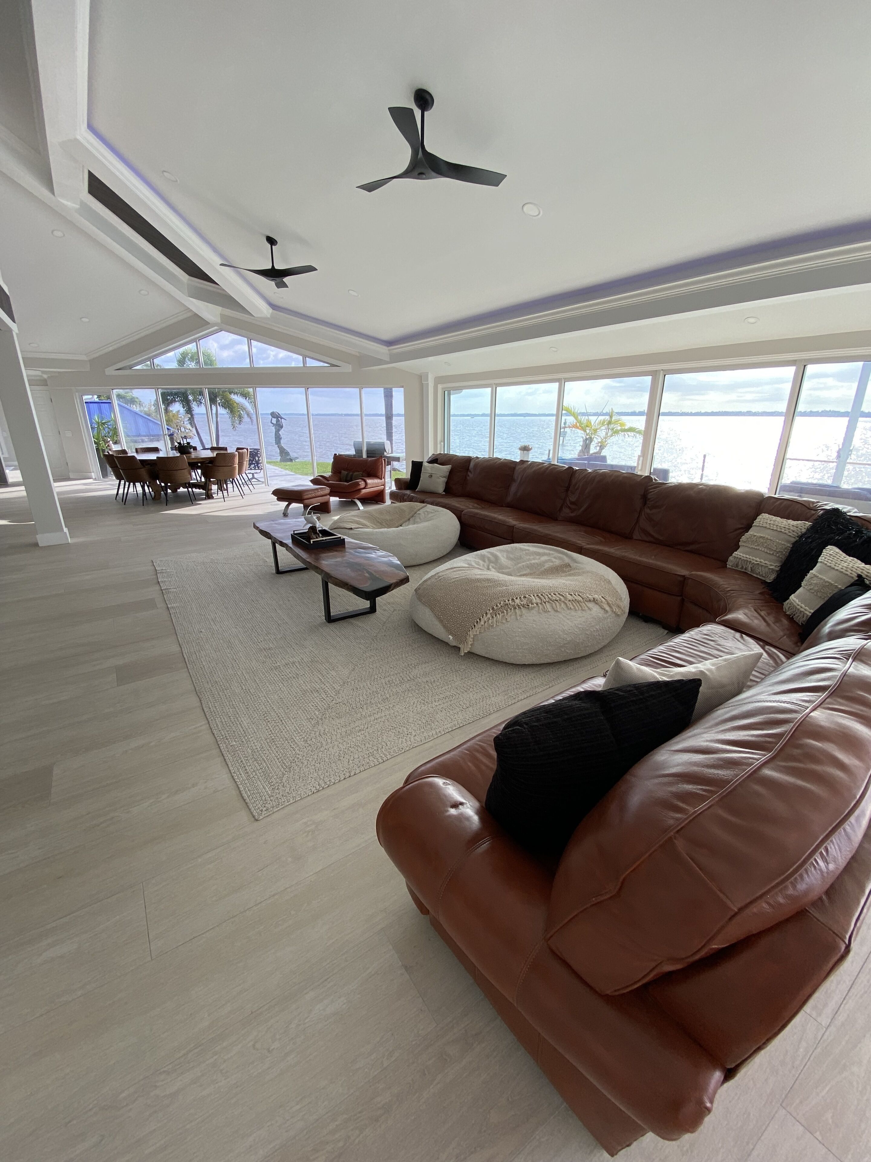 Uma sala de estar ampla com um sofá de couro marrom grande, uma mesa de centro e uma área de jantar com mesa e cadeiras. Há duas ventoinhas de teto e janelas amplas com vista para a água.