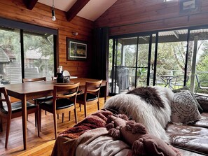 A cozy room with a wooden dining table and chairs, a sofa with a fluffy throw, and large windows offering a view of the outdoors.
