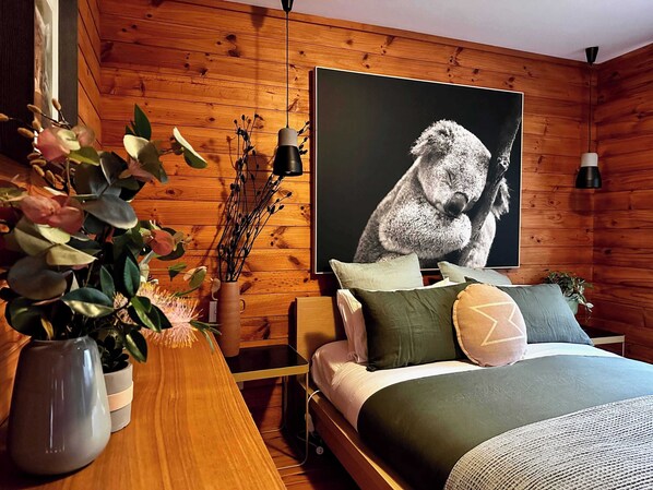 A cozy bedroom with wooden walls, a large bed with multiple pillows, a bedside table with a vase of flowers, and a wall-mounted picture.