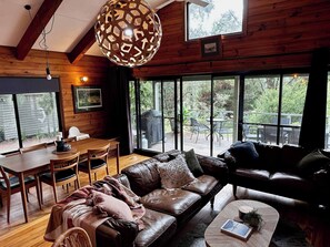 A cozy living room with a wooden ceiling, a large window overlooking a deck, and a dining area with a table and chairs.