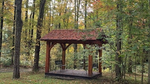 Un kiosque en bois avec un toit en tuiles, entouré d’arbres et de feuilles mortes.