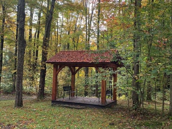 A wooden gazebo with a tiled roof, surrounded by trees and fallen leaves.