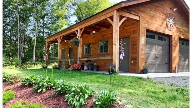 Une cabane en bois dotée d’un porche abrité, d’un garage et d’un jardin bien entretenu où poussent différentes espèces de plantes.