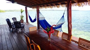 Una terraza de madera con una mesa y sillas, una hamaca azul, y vista al agua.