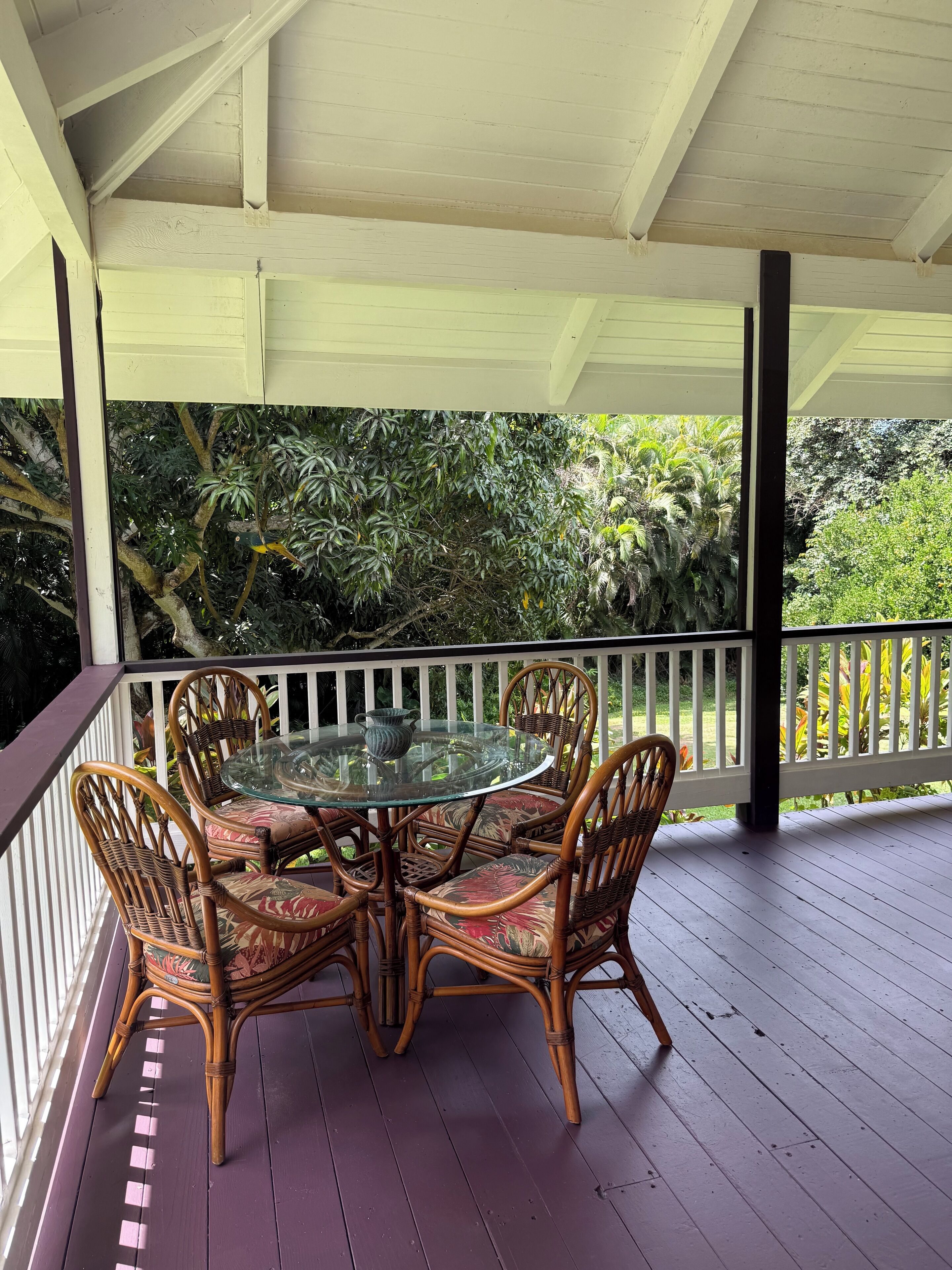 A covered outdoor seating area with a glass table and wicker chairs, surrounded by a white railing and lush greenery.