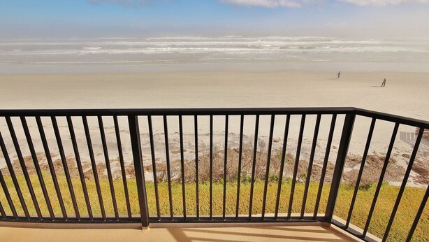 Vue sur la plage depuis un balcon doté d’une rambarde en métal, avec une plage de sable et des vagues de l’océan.