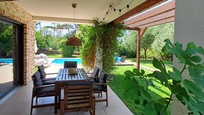 A covered patio area with a wooden table and chairs, a hanging light fixture, and a view of a swimming pool and greenery. - Peaceful house with lots of privacy near Umag. Sea view. (Buje)