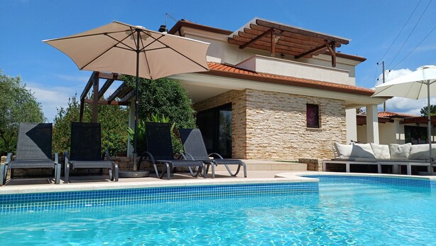 A swimming pool with lounge chairs and umbrellas, a stone building with a tiled roof, and a clear blue sky. - Peaceful house with lots of privacy near Umag. Sea view. (Buje)