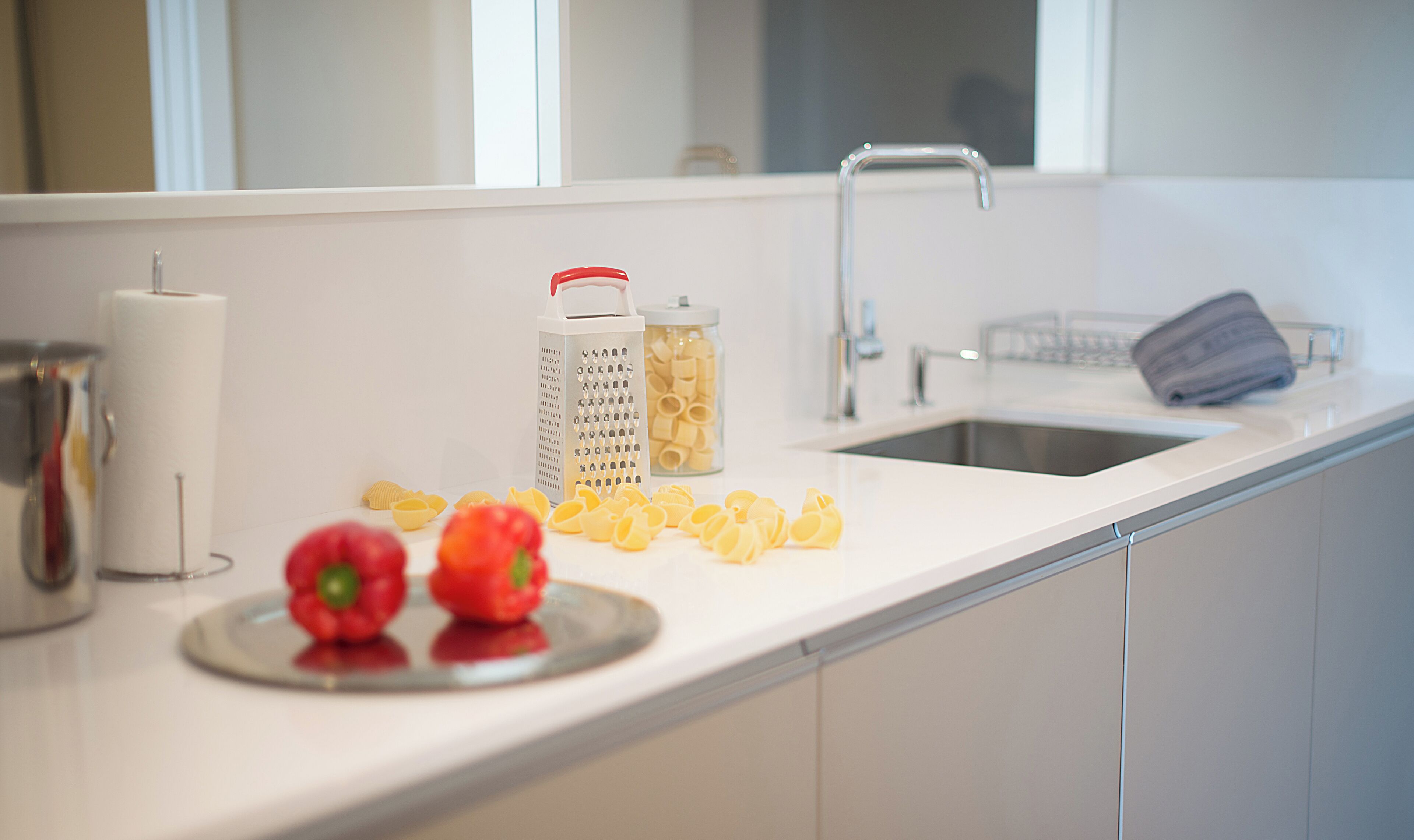 Una encimera de cocina con un lavabo, un rallador, un tarro de pasta y dos pimientos rojos.