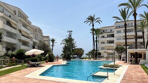 Une piscine dotée d’une terrasse carrelée, entourée de palmiers et bordée d’un immeuble à plusieurs étages.