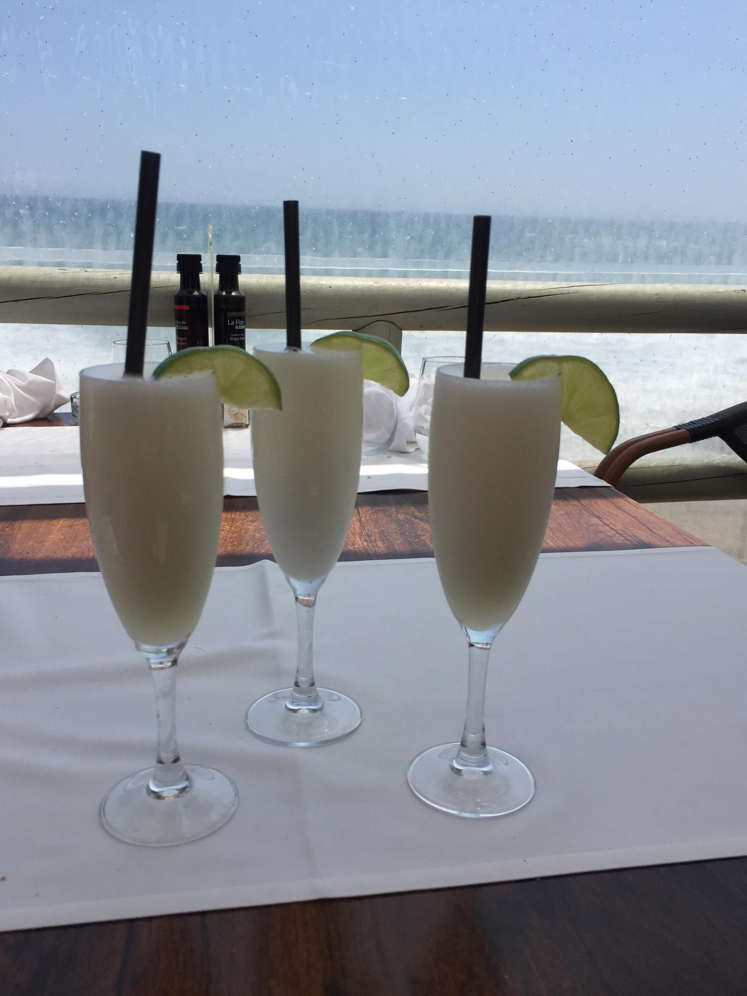 Trois verres d’une boisson blanche, décorés de rondelles de citron vert, sont posés sur une table près d’une fenêtre donnant sur la plage.