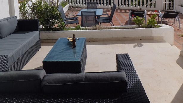 Une terrasse extérieure avec un canapé, une table et des chaises. On peut admirer l’océan et, au loin, des palmiers.