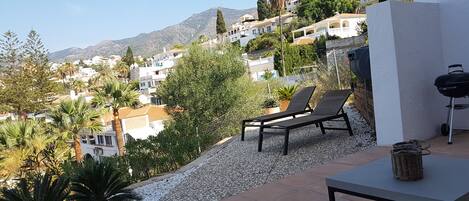 A patio with a table and two lounge chairs, overlooking a hillside of houses and a mountain.