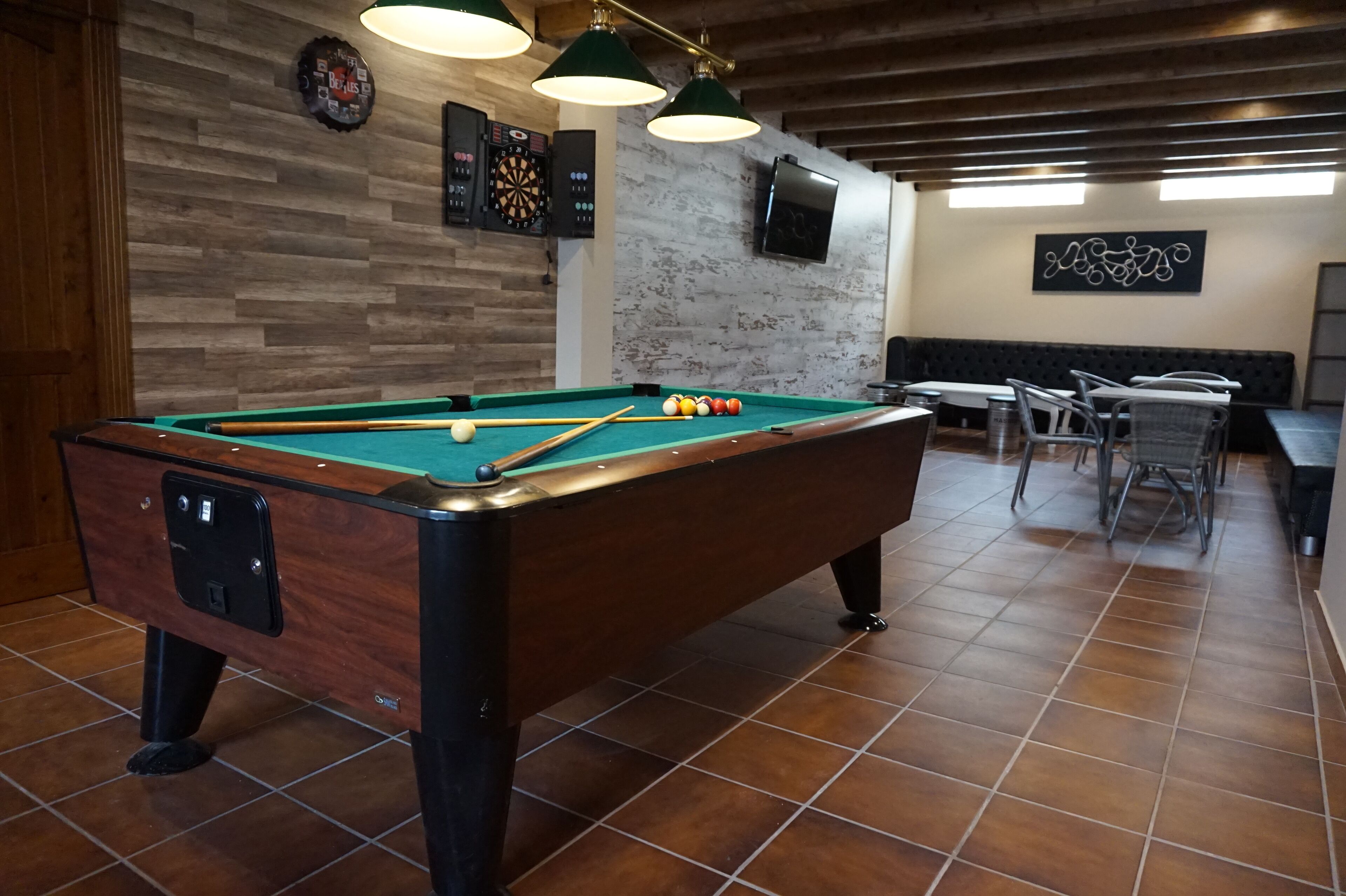 A pool table with balls, a dartboard, and a billiards table in a room with wooden walls and tiled flooring.