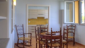 Zona pranzo con tavolo e sedie in legno, cucina sullo sfondo e una finestra con persiane.