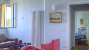 Un soggiorno con un divano grigio, un pouf rosa e un tavolino con un vaso e delle candele. Si nota un termosifone, una finestra e una porta che conduce in un'altra stanza.