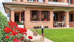 Una casa de ladrillo de dos pisos con un porche cubierto, un pequeño jardín con flores rojas y un césped.