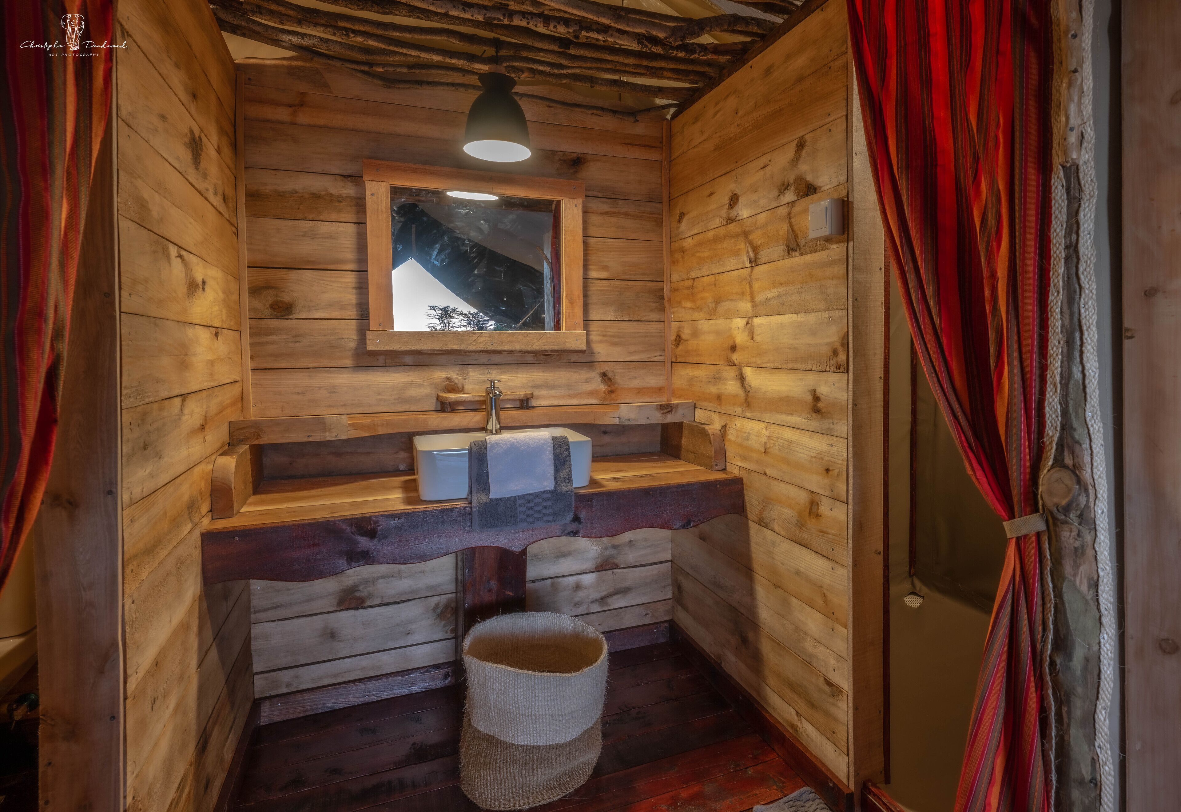 A wooden cabin bathroom with a window, wooden bench, and a hanging light fixture.