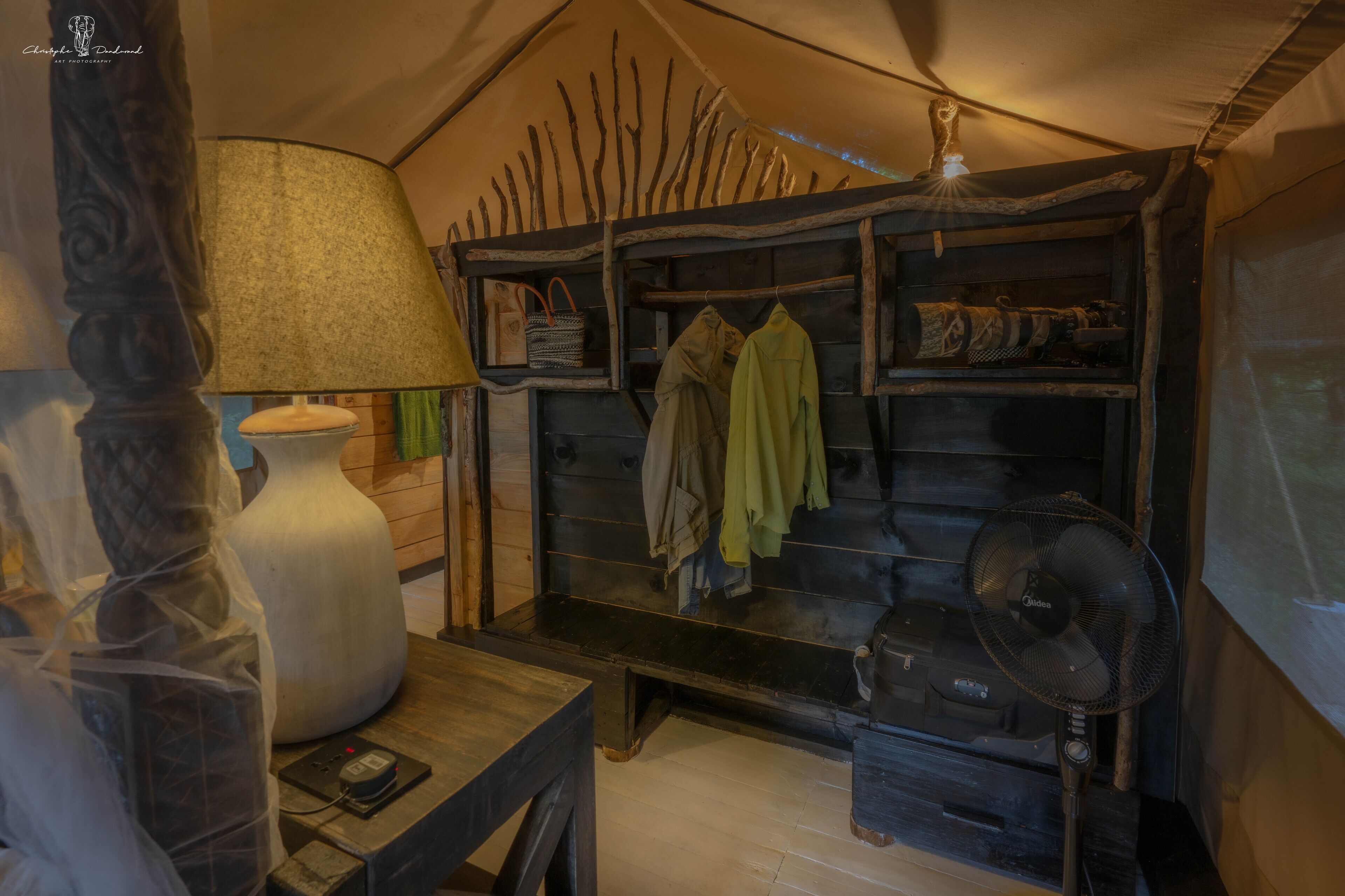 A cozy room with a large lamp, a wooden shelf with clothes, and a fan.