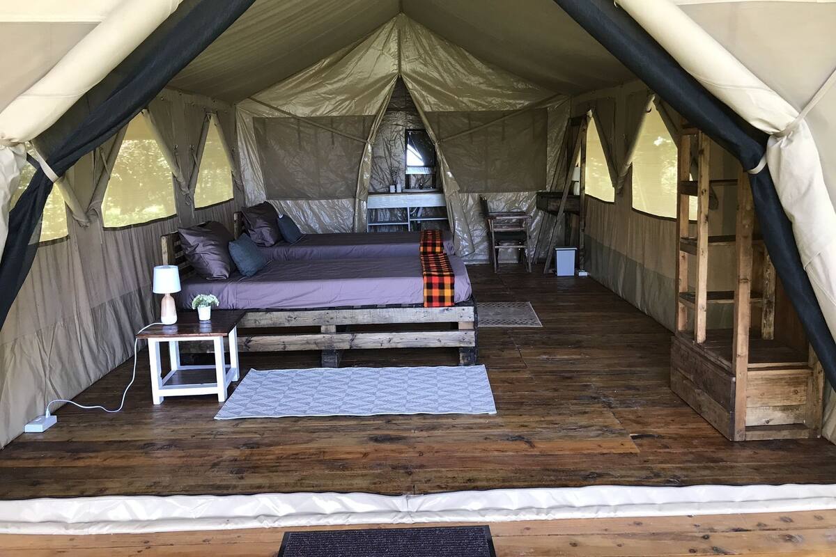 Una tienda de campaña con una cama, una mesita con una lámpara y una silla.