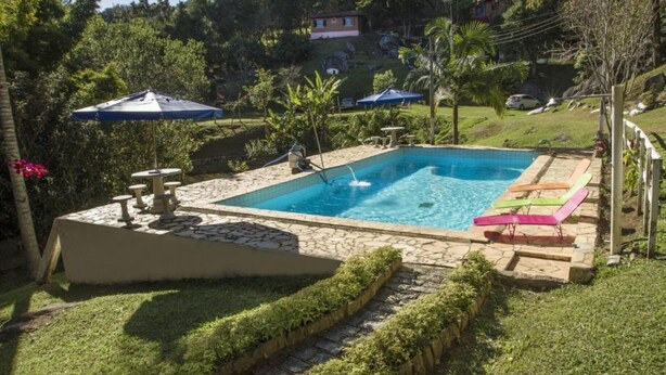 A rectangular swimming pool with a stone border, surrounded by a grassy area with a few palm trees and a stone bench. - Pousada Céu de Luz - Próximo a Monte Verde (Itapeva)