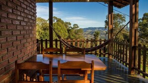 Una mesa y sillas de madera bajo un porche cubierto con un muro de ladrillo, una hamaca y vistas a colinas y árboles.