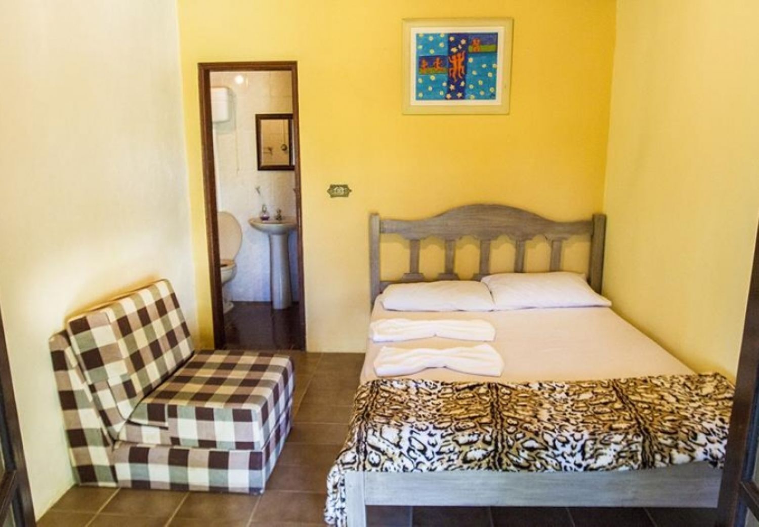 A small room with a bed, a checkered bench, a sink, and a framed artwork on the wall.