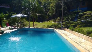 A swimming pool with a stone border, surrounded by palm trees and a garden. - Pousada Céu de Luz - Próximo a Monte Verde (Itapeva)