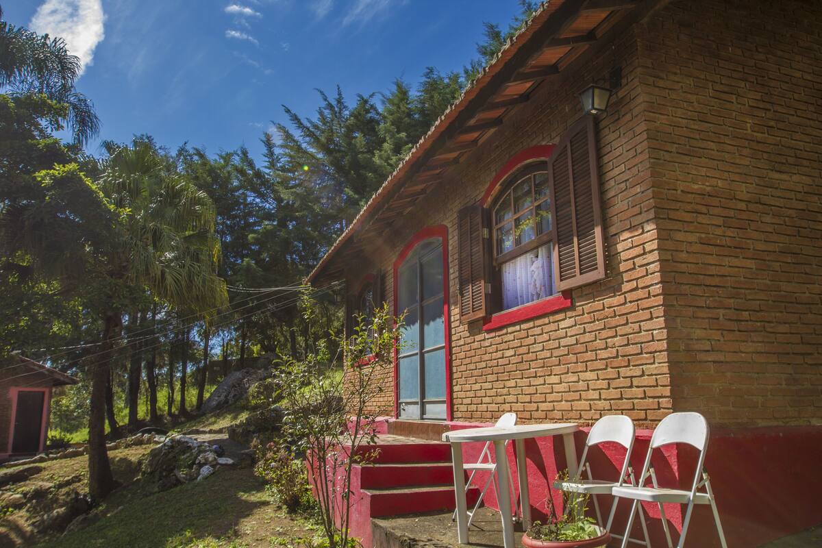 A rustic brick building with a red exterior and a small porch area. There are white chairs and a table on the porch.