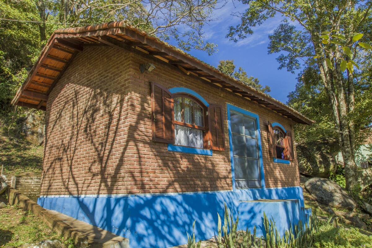 A rustic brick house with a blue base and wooden roof, surrounded by trees and vegetation.