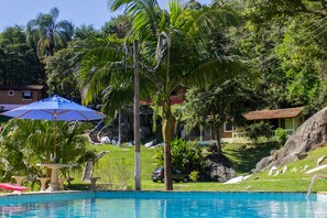 A swimming pool with a blue umbrella, palm trees, and a house in the background. - Pousada Céu de Luz - Próximo a Monte Verde (Itapeva)