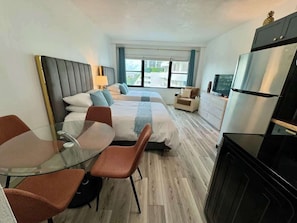 Une chambre d’hôtel avec un lit, une table en verre et des chaises, une télévision, un réfrigérateur et une vue sur les bâtiments environnants.
