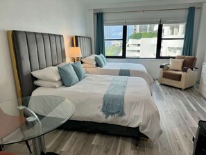 Une chambre d’hôtel avec deux lits, une table en verre, une chaise et une vue sur les immeubles par la fenêtre.