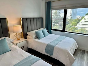 Habitación de hotel con dos camas, una mesita de noche con lámpara, una ventana con vistas a edificios y árboles, y un diseño moderno.