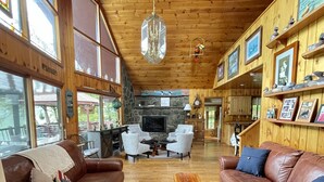 A spacious living room with leather sofas, a central wooden coffee table, and a stone fireplace. Large windows allow natural light to fill the space.