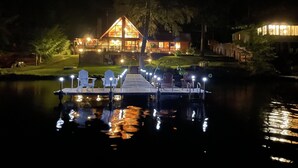 A lakeside lodge with a dock, chairs, and a well-lit building.
