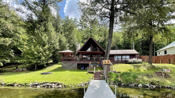 A lodge with a wooden deck leading to a dock over a pond, surrounded by lush greenery and trees.