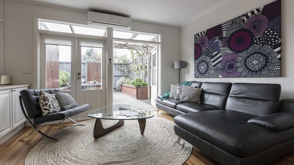 Relax in style with a comfortable sofa, armchair, and abstract artwork, surrounded by lush greenery through the sliding glass door.