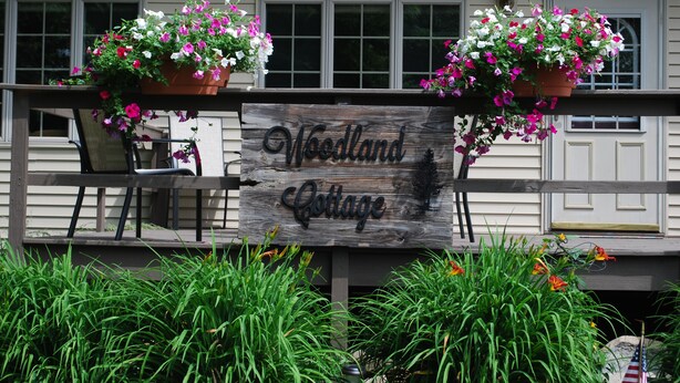 Un panneau en bois sur lequel est inscrit « Woodland Cottage », entouré de plantes en pot et de verdure.