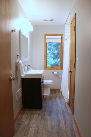 A narrow bathroom with a wooden door, a white sink, a toilet, and a window with a view of trees. - Cozy Cottage near Schwarz Supper Club, Road America, and Whistling Straights!! (New Holstein)