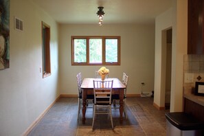 A dining area with a wooden table, four chairs, a vase with flowers, and a window with a view of trees. - Cozy Cottage near Schwarz Supper Club, Road America, and Whistling Straights!! (New Holstein)