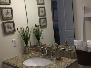 A bathroom with a granite countertop, a white sink, a mirror, framed artwork on the wall, and a door leading to another room. - Spectacular Views From Beachfront Condo (Hampton)