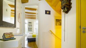 A hallway with a wooden floor, a bed in the background, a yellow door, and a clock on the wall.