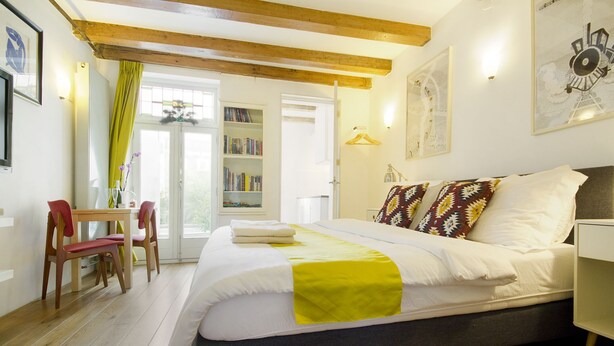 A bedroom with a bed, a desk, chairs, a bookshelf, and a door leading to a balcony.