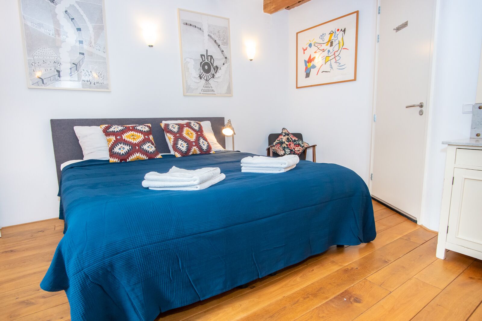 A neatly made bed with blue bedding, a chair with a floral pillow, and artwork on the walls.