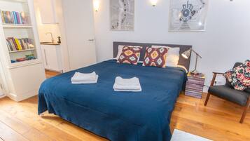 Una cama bien hecha con una colcha azul y dos toallas blancas dobladas, una estantería con libros, un escritorio con una lámpara, y dos obras de arte enmarcadas en la pared.