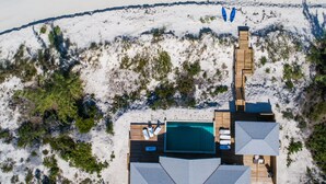 Vue aérienne d’un complexe hôtelier situé en bord de mer, avec une grande piscine, une terrasse en bois et une plage.